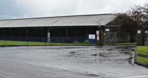 A single storey warehouse surrounded by a fence and showing a driveway into parking adjacent to the warehouse